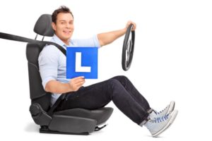 Young man holding a steering wheel and an L-sign seated on a car seat isolated on white background
